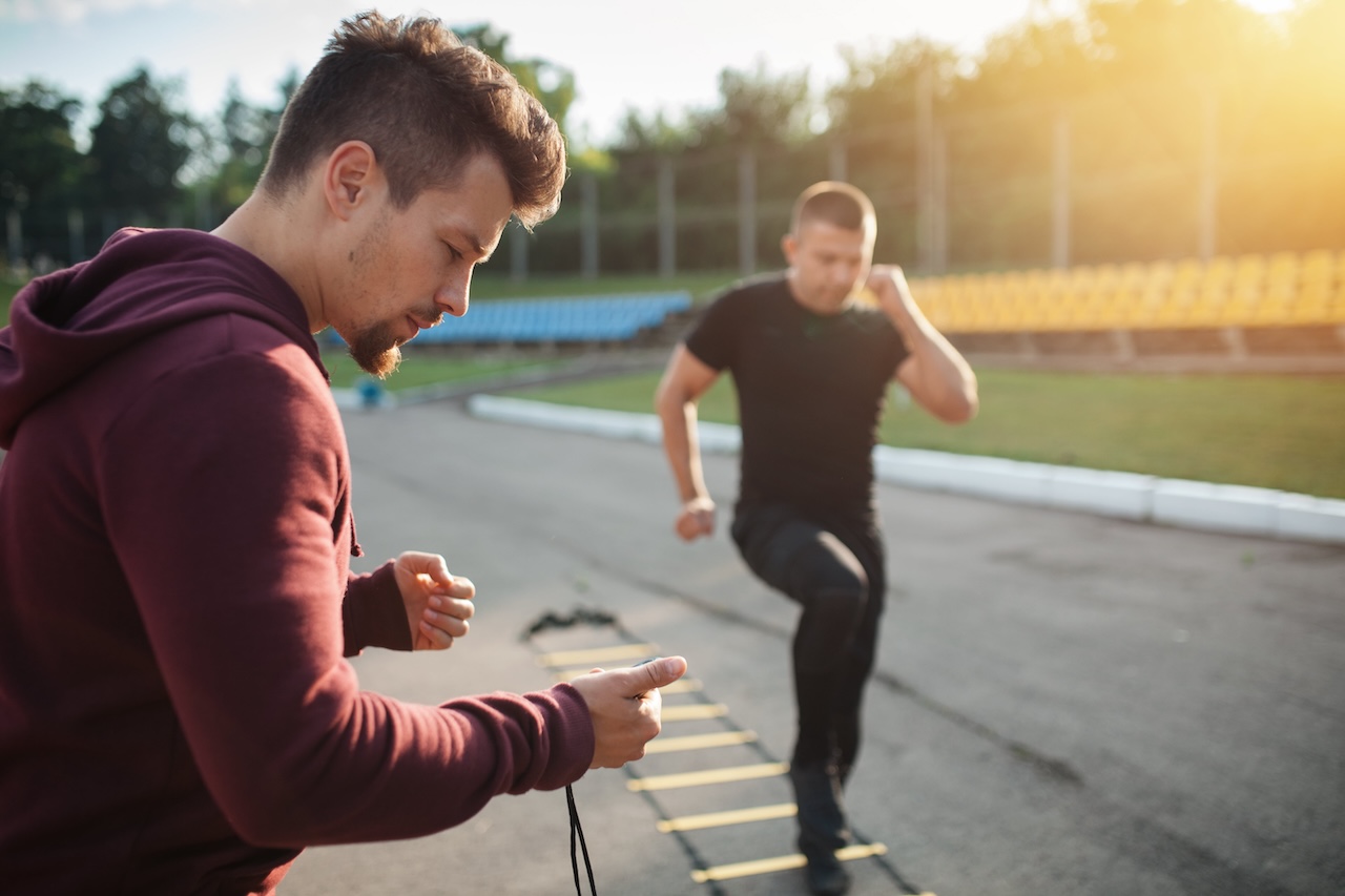 A sports coach looking at his stopwatch and a person exercising outdoors.