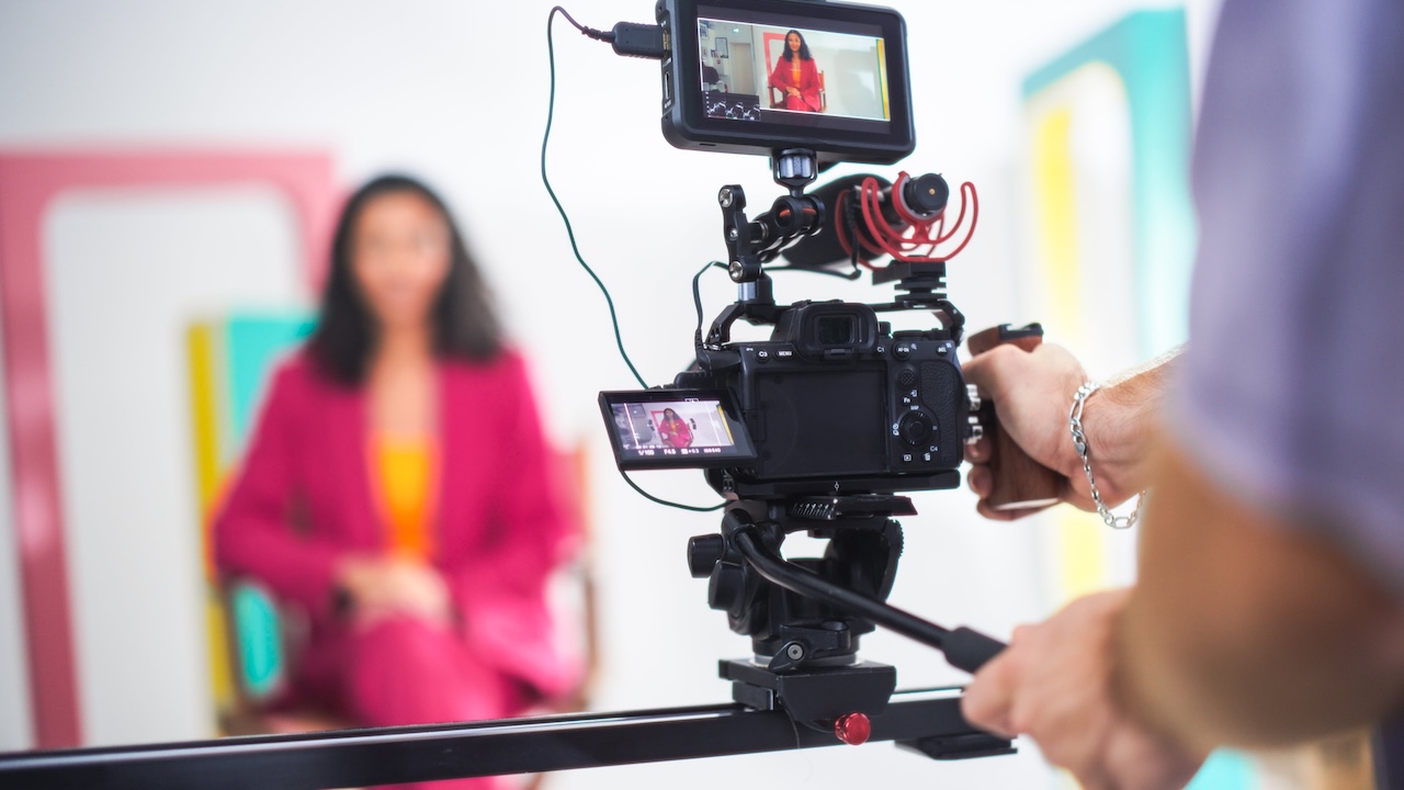 Film set, with a person using professional filming equipment to film a host speaking to camera.